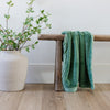 A Luxury Receiving Seagrass (dusty green) Colored Lush Saranoni Blanket draped over a wood bench. The soft blanket is a small blanket and a baby blanket or toddler blanket.