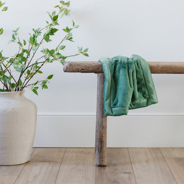 A Mini Seagrass (dusty green) Colored Lush Saranoni Blanket lays over a wood bench. The soft blanket is a small blanket and a baby blanket or toddler blanket.