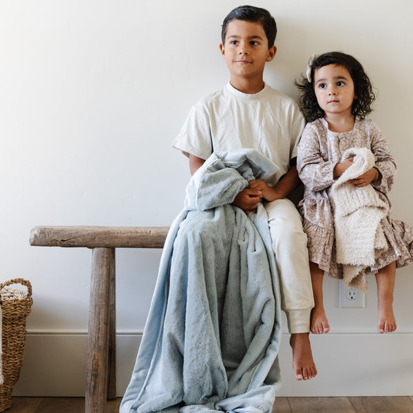 A little boy holds a Toddler Chambray (cloud-like light blue) Colored Lush Luxury Saranoni Blanket. The soft blanket is a baby blanket or toddler blanket. He is sitting by his younger sister who is holding a Mini Bamboni Saranoni Blanket.