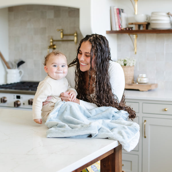 A mother holds her baby boy who has a Luxury Receiving Chambray (cloud-like light blue) Colored Lush Saranoni Blanket draped over his lap. The soft blanket is a small blanket and a baby blanket or toddler blanket.