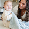 A mother holds her baby boy who has a Luxury Receiving Chambray (cloud-like light blue) Colored Lush Saranoni Blanket draped over his lap. The soft blanket is a small blanket and a baby blanket or toddler blanket.