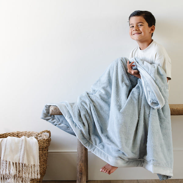 A little boy holds a Toddler Chambray (cloud-like light blue) Colored Lush Luxury Saranoni Blanket. The soft blanket is a baby blanket or toddler blanket.