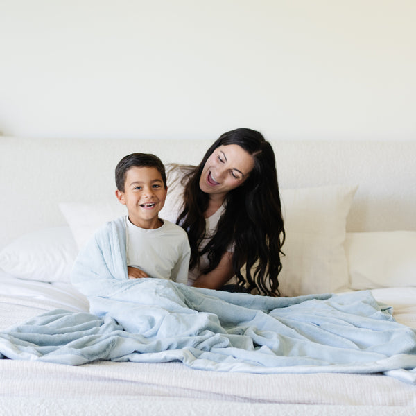 A mother and her son sit under a Luxury Extra Large Oversized Throw Chambray (cloud-like light blue) Colored Lush Saranoni Blanket. The soft blanket is an extra large throw blanket and has a soft faux fur feeling.