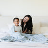 A mother and her son sit under a Luxury Extra Large Oversized Throw Chambray (cloud-like light blue) Colored Lush Saranoni Blanket. The soft blanket is an extra large throw blanket and has a soft faux fur feeling.