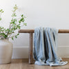 A Chambray (cloud-like light blue) Colored Lush Saranoni Blanket lays over a wood bench. The soft blanket is a small blanket and a baby blanket or toddler blanket.