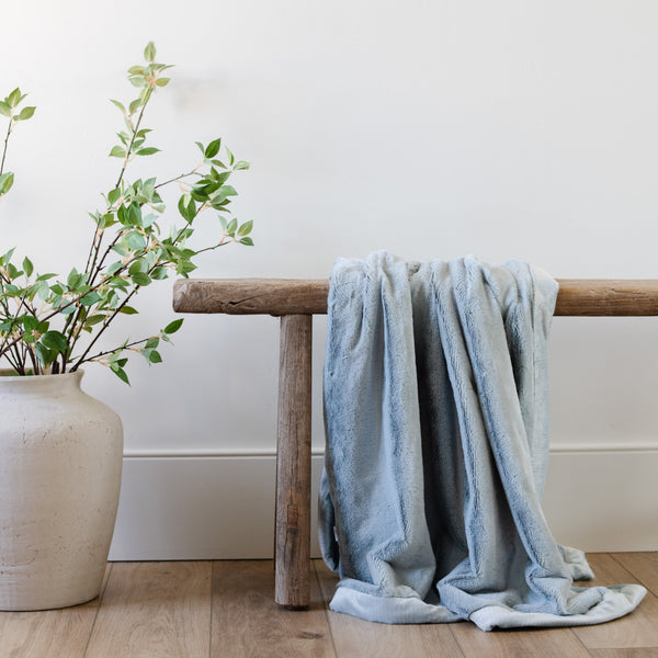 A Luxury Receiving Chambray (cloud-like light blue) Colored Lush Saranoni Blanket draped over a wooden bench. The soft blanket is a small blanket and a baby blanket or toddler blanket.