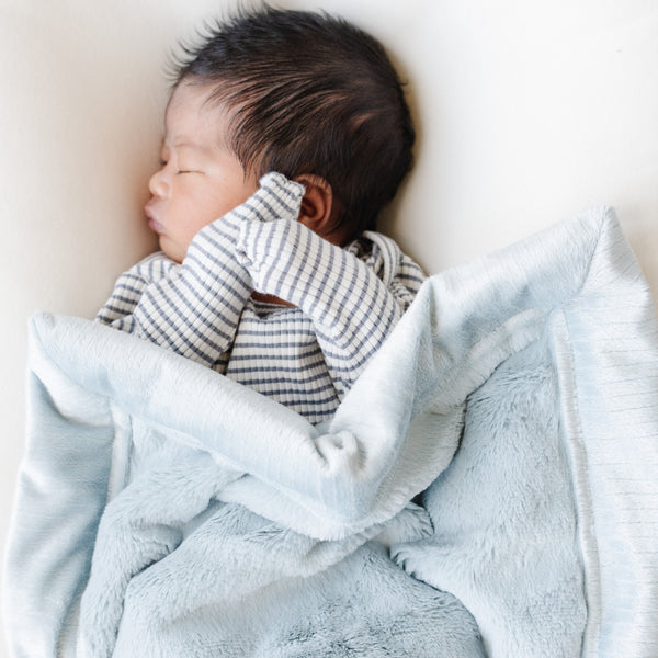 A newborn baby boy lays under a Mini Chambray (cloud-like light blue) Colored Lush Saranoni Blanket. The soft blanket is a small blanket and a baby blanket or toddler blanket.