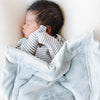 A newborn baby boy sleeps under a Luxury Receiving Chambray (cloud-like light blue) Colored Lush Saranoni Blanket that is draped over him. The soft blanket is a small blanket and a baby blanket or toddler blanket.