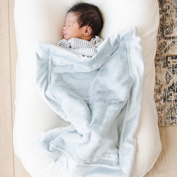 A newborn baby boy lays under a Mini Chambray (cloud-like light blue) Colored Lush Saranoni Blanket. The soft blanket is a small blanket and a baby blanket or toddler blanket.