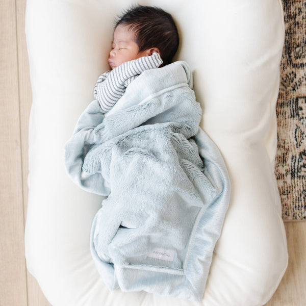 A newborn baby boy lays under a Mini Chambray (cloud-like light blue) Colored Lush Saranoni Blanket. The soft blanket is a small blanket and a baby blanket or toddler blanket.