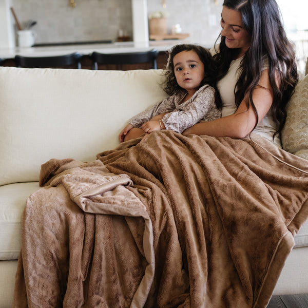 A mother holds her daughter while sitting under a Luxury Extra Large Oversized Throw Hazelnut Colored Lush Saranoni Blanket. The soft blanket is an extra large throw blanket and has a soft faux fur feeling.