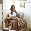 A woman reads a book while sitting under a Luxury Extra Large Oversized Throw Hazelnut Colored Lush Saranoni Blanket. The soft blanket is an extra large throw blanket and has a soft faux fur feeling.