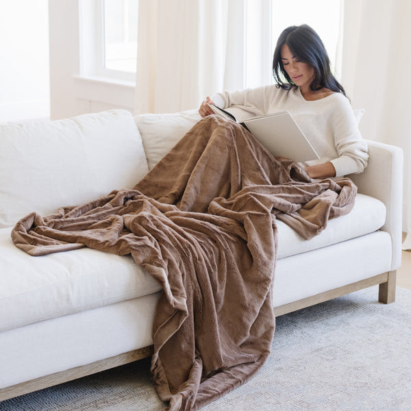 Woman reading on a white couch with a Hazelnut Lush XL Saranoni blanket, designed for oversized comfort and featuring a buttery soft, plush texture. This luxurious Saranoni blanket is perfect for cozy evenings, reading, or home decor. The Saranoni blanket adds warmth, style, and unmatched softness, making it an ideal gift or essential for ultimate relaxation.