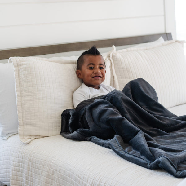 A little boy sits under a Luxury Toddler Charcoal Colored Lush Saranoni Blanket. The soft blanket is a toddler blanket or baby blanket.