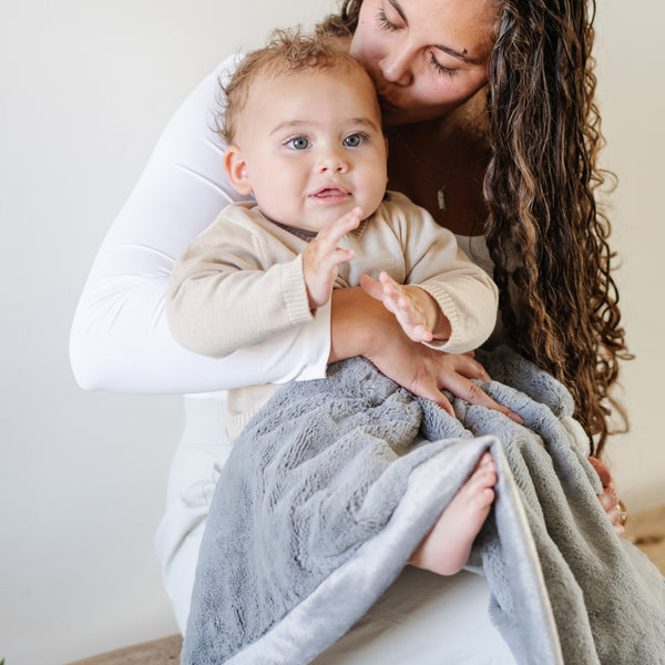 A mother holds her baby boy and a Luxury Receiving Gray Colored Lush Saranoni Blanket over her baby boy's lap. The soft blanket is a small blanket and a baby blanket or toddler blanket.