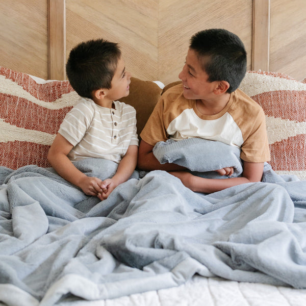 Two brothers sit while holding a Luxury Extra Large Oversized Throw Gray Colored Lush Saranoni Blanket. The soft blanket is an extra large throw blanket and has a soft faux fur feeling.