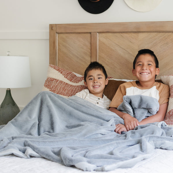 Two brothers sit while holding a Luxury Extra Large Oversized Throw Gray Colored Lush Saranoni Blanket. The soft blanket is an extra large throw blanket and has a soft faux fur feeling.