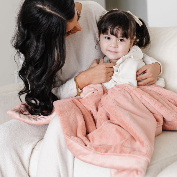 A mother and her little girl sit under a Luxury Receiving Blossom (light peachy pink) Colored Lush Saranoni Blanket. The soft blanket is a small blanket and a baby blanket or toddler blanket.