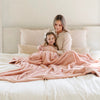 A mother and her daughter sit under a Luxury Extra Large Oversized Throw Blossom (light peachy pink) Colored Lush Saranoni Blanket. The soft blanket is an extra large throw blanket and has a soft faux fur feeling.