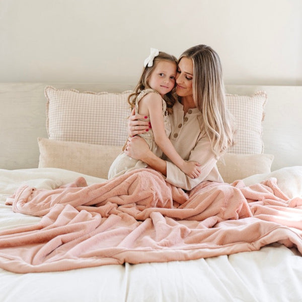 A mother and her daughter sit under a Luxury Extra Large Oversized Throw Blossom (light peachy pink) Colored Lush Saranoni Blanket. The soft blanket is an extra large throw blanket and has a soft faux fur feeling.