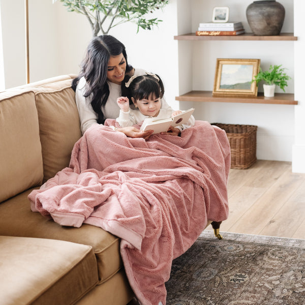 A mother and daughter read a story while holding a Luxury Extra Large Oversized Throw Ballet Slipper (light pink) Colored Lush Saranoni Blanket. The soft blanket is an extra large throw blanket and has a soft faux fur feeling.