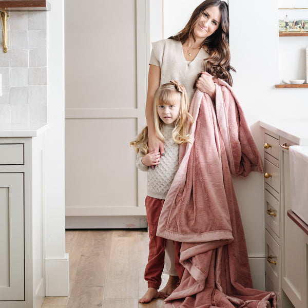 A mother holds her daughter while holding a Luxury Extra Large Oversized Throw Ballet Slipper (light pink) Colored Lush Saranoni Blanket. The soft blanket is an extra large throw blanket and has a soft faux fur feeling.