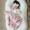 A Little Baby Girl with a white bow and white outfit and a Mini Saranoni Lush Blanket that is light pink. The specific color is called Ballet Slipper.