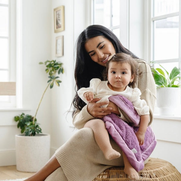 A mother wearing a tan outfit holds her little girl that is wearing a white outfit. The mother has a Mini Saranoni Lush Blanket draped over her daughter. The blanket is a lighter purple. The specific color is called Fairy Wings.