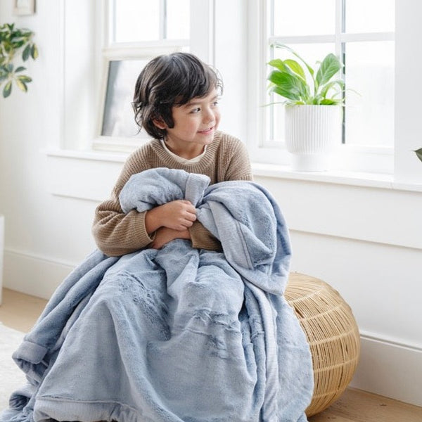 A little boy sits under a Luxury Receiving Storm Cloud (dusty blue gray) Colored Lush Saranoni Blanket. The soft blanket is a small blanket and a baby blanket or toddler blanket.