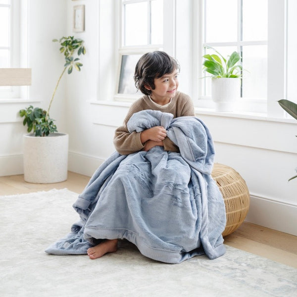 A little boy sits under a Luxury Toddler Storm Cloud (dusty blue gray) Colored Lush Saranoni Blanket. The soft blanket is a toddler blanket or baby blanket.