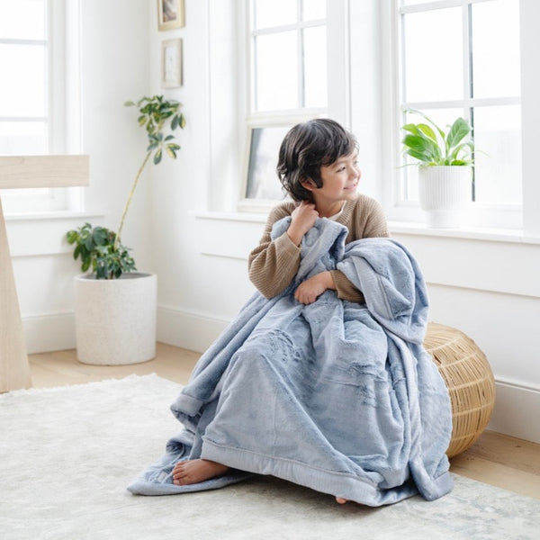 A little boy sits under a Luxury Toddler Storm Cloud (dusty blue gray) Colored Lush Saranoni Blanket. The soft blanket is a toddler blanket or baby blanket.