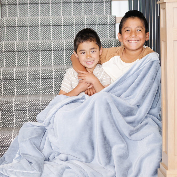 Two little boys who are brothers sit under a Luxury Toddler Storm Cloud (dusty blue gray) Colored Lush Saranoni Blanket. The soft blanket is a toddler blanket or baby blanket.