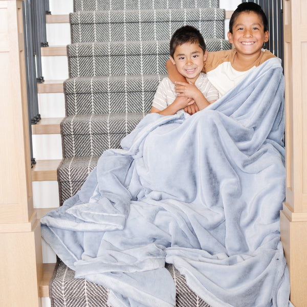 Two little boys who are brothers sit under a Storm Cloud (light blue/gray) Lush XL Saranoni blanket, designed for oversized comfort and featuring a buttery soft, plush texture. This luxurious Saranoni blanket is perfect for cozy evenings, reading, or home decor. The Saranoni blanket adds warmth, style, and unmatched softness, making it an ideal gift or essential for ultimate relaxation.