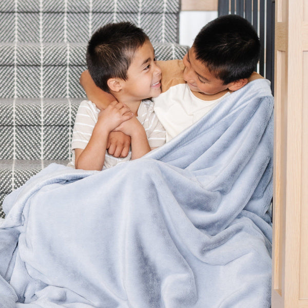 Two little boys who are brothers sit under a Luxury Toddler Storm Cloud (dusty blue gray) Colored Lush Saranoni Blanket. The soft blanket is a toddler blanket or baby blanket.