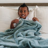 A little boy sits under a Luxury Toddler Eucalyptus Colored Lush Saranoni Blanket. The soft blanket is a toddler blanket or baby blanket.