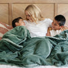 A mother and her two sons sit under a Luxury Extra Large Oversized Throw Hunter Green Colored Lush Saranoni Blanket. The soft blanket is an extra large throw blanket and has a soft faux fur feeling.