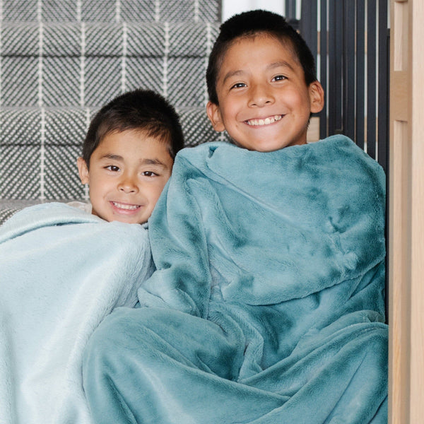 A little boy holds a Toddler Mineral Blue Colored Lush Luxury Saranoni Blanket. The soft blanket is a small blanket and a baby blanket or toddler blanket. His brother sitting to the left of him has a Dew Colored Lush Saranoni Blanket.