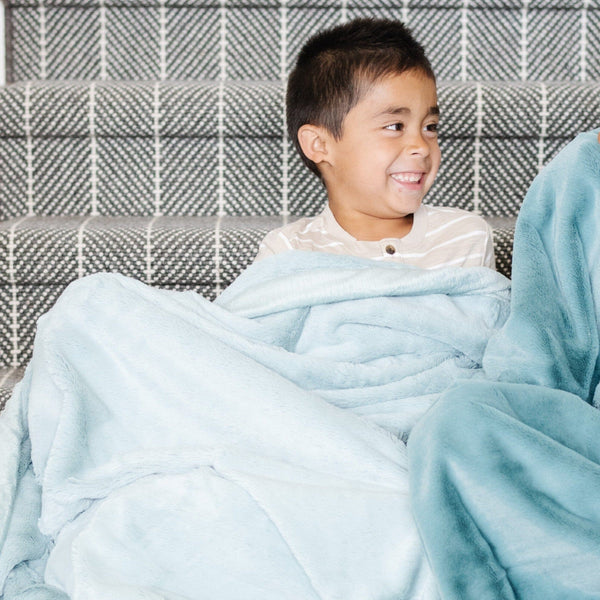 A little boy sits under a Luxury Toddler Dew (light blue) Colored Lush Saranoni Blanket. The soft blanket is a toddler blanket or baby blanket.