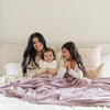 A mother and her two daughters sit under a Luxury Extra Large Oversized Throw Bloom (dusty purple) Colored Lush Saranoni Blanket. The soft blanket is an extra large throw blanket and has a soft faux fur feeling.