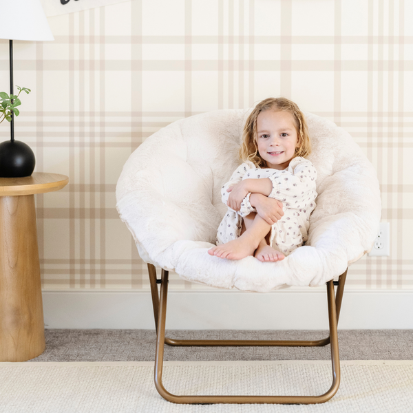 A little girl lounging in a cozy cream Saranoni faux fur saucer chair, featuring a soft, removable faux fur cover made from the same luxurious fabric as Saranoni’s blankets. This stylish and foldable saucer chair offers a safe, comfortable space for kids to relax, read, or play. Perfect for adding warmth and texture to any room, it’s a must-have for creating a kid-friendly lounging spot.