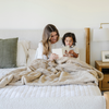 A mother sits reading a book with her son. They have a khaki patterned faux fur blanket on their lap. The soft blanket is a Saranoni blanket and a luxury blanket. 