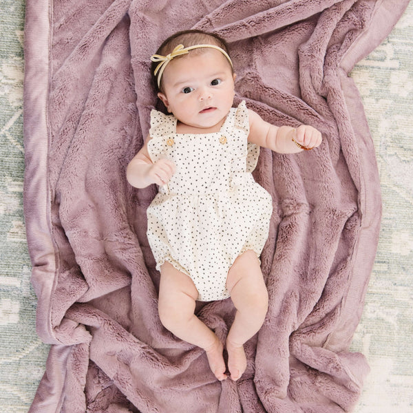 A baby girl lays on a Receiving Bloom (dusty purple) Colored Lush Saranoni Blanket. The soft blanket is a small blanket and a baby blanket or toddler blanket.