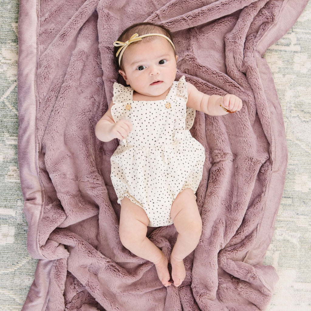 A baby girl lays on a Receiving Bloom (dusty purple) Colored Lush Saranoni Blanket. The soft blanket is a small blanket and a baby blanket or toddler blanket.