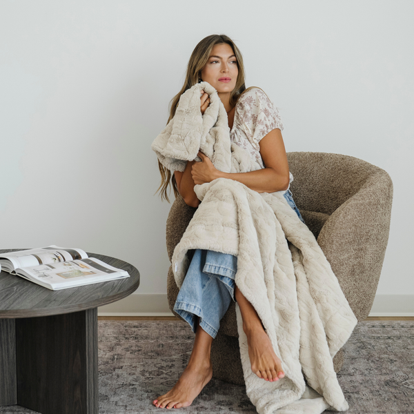 Woman sits in chair wrapped in a Buff cozy faux fur throw blanket. 