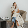 Woman sits in chair wrapped in a Buff cozy faux fur throw blanket. 