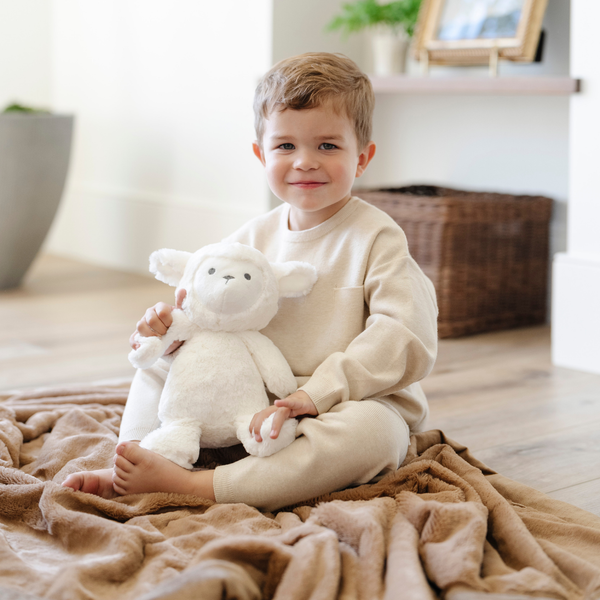 A little boy holds a white lamb stuffed animal from Saranoni while sitting on a hazelnut colored lush blanket. The soft blanket is a Saranoni blanket.