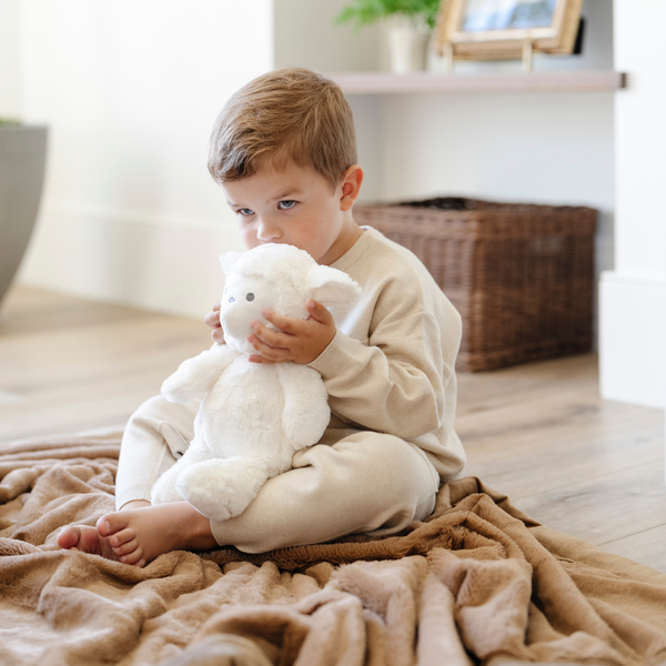 A little boy holds a white lamb stuffed animal from Saranoni while sitting on a hazelnut colored lush blanket. The soft blanket is a Saranoni blanket.