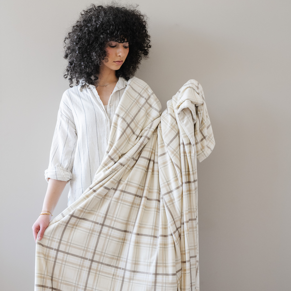 Woman stands holding a neutral plaid minky stretch XL blanket. 