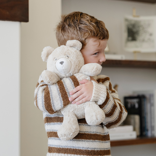 A little boy hugs a light tan bear stuffed animal. The bear stuffed animal is from Saranoni. 