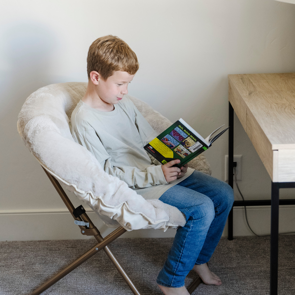 A little boy reads a book while sitting in a cozy light tan Saranoni faux fur saucer chair, featuring a soft, removable faux fur cover made from the same luxurious fabric as Saranoni’s blankets. This stylish and foldable saucer chair offers a safe, comfortable space for kids to relax, read, or play. Perfect for adding warmth and texture to any room, it’s a must-have for creating a kid-friendly lounging spot.